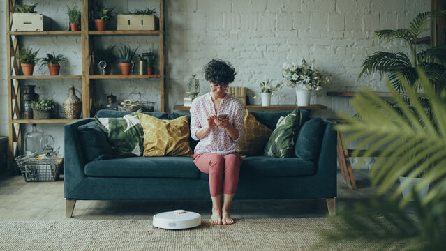 Cheerful young lady is turning on cool robotic hoover and enjoying automatic cleaning sitting on sofa and relaxing with smartphone. Modern technology and youth concept.