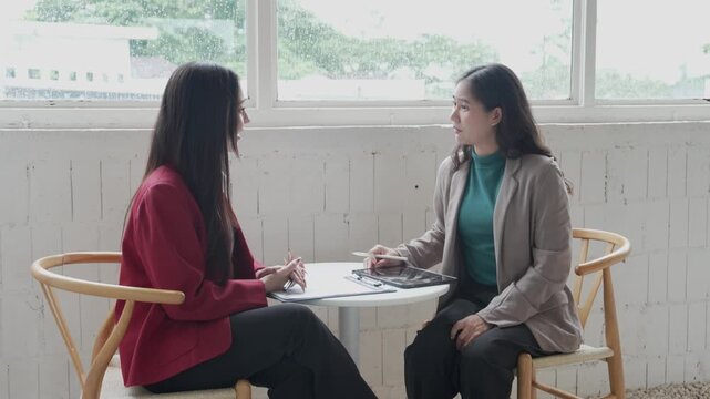 Two Asian businesswomen sitting at a small office table, discussing work details using a digital tablet and taking notes on paper documents during a business meeting near a large window.