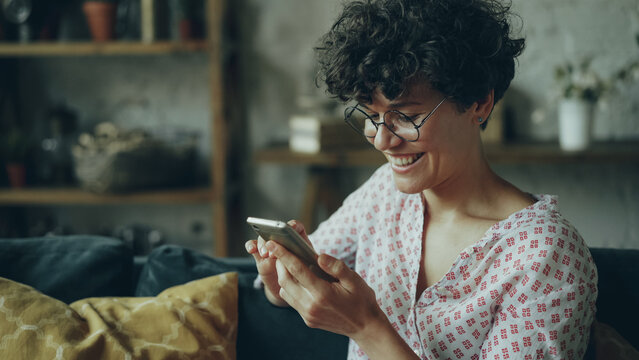 Happy girl is using modern smartphone looking at screen and laughing texting friends and checking social media accounts in internet sitting on sofa at home.