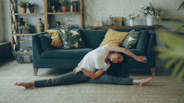 Slender Asian lady with long hair is doing sports at home stretching body, arms and legs in seated position on floor enjoying physical activity and healthy lifestyle. Sport and millennials concept.