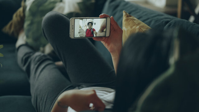 Young woman with long dark hair is making online video call holding smartphone and gesturing talking to male friend lying on sofa in house. Youth and informal communication concept.
