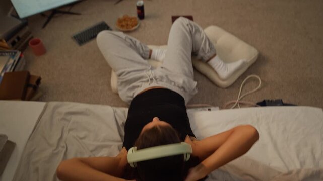 Multiethnic young woman typing at monitors, leaning back on bed wearing headphones, looking up. Visual suited for remote work fatigue, mental wellbeing, or music app marketing