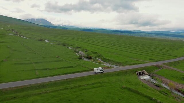 A white camper rides on an empty asphalt road through a vast green landscape with snow-capped mountains in the background, embodying the concept of freedom, travel and adventure vacation in Georgia