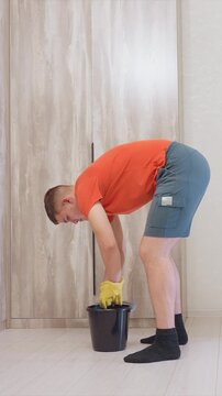 Man in orange shirt scrubs wooden floors with mops while kneeling using bright yellow gloves. setting is clean indoor space with light colored wood panel walls and flooring