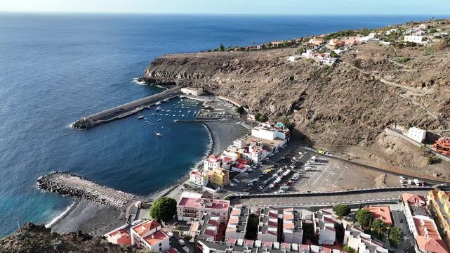Aerial pull back drone shot revealing the scenic coastal town Playa de Santiago on La Gomera, Canary Islands. The rugged landscape, a small harbor, beach, and houses are seen near Roque de la Cruz.