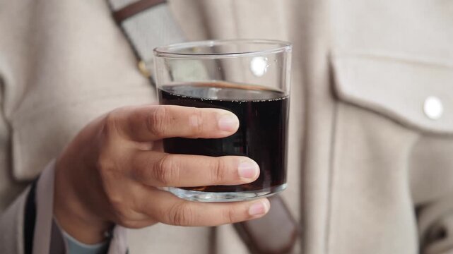 Woman holding glass of carbonated dark soda beverage in her hand outdoors close up shot.