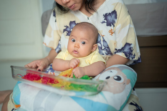 Asian Baby Looking at Camera while Grabbing Colorful Spaghetti Noodles in Mother Lap During Sensory Play