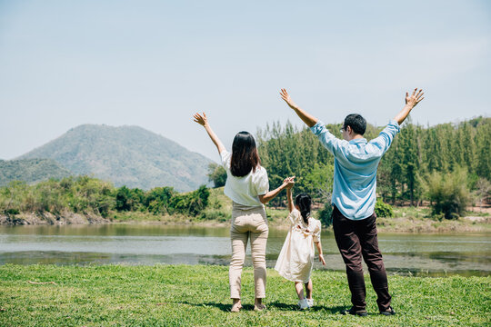 Asian family father mother and daughter stand front lake with raised hands sunny day in park, lifestyle parents and child kid enjoying outdoors together in nature on summer travel
