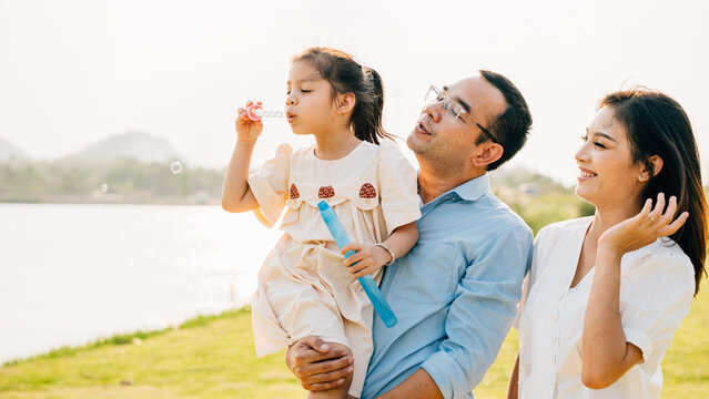 Asian family having fun playing together on spring meadow field grass in park, Happy parents father carrying daughter and a child blowing soap bubbles enjoying outdoors together