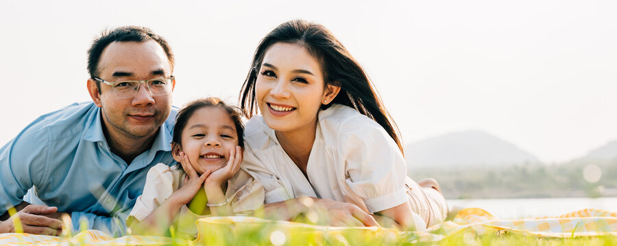 Asian family father mother and daughter lying on blanket spring meadow field grass in park cheerful sweet kid girl, parents and child picnic enjoying outdoors sunset time together