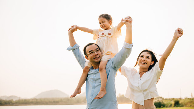 Asian family child girl piggyback riding back on father raising hands like flying outdoor nature park with mother, Happy parent daughter having fun together