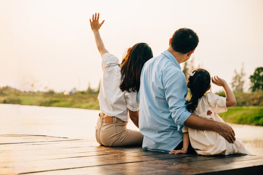 Asian family of three a girl child are sitting on a wooden dock by a body of water. Happy parent father mother and daughter kid they are all smiling