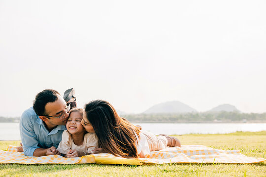Asian family father mother and daughter lying on blanket spring meadow field grass in park cheerful sweet kid girl, parents and child picnic enjoying outdoors sunset time together