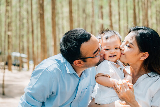 Asian family parents embracing in a lush green forest nature kissing child, happy father mother have picnic kiss his daughter together