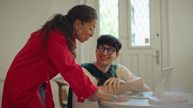 Woman mentors a man at a laptop in a bright workspace, showing training, teamwork, business learning, coaching, and intergenerational collaboration during focused one-on-one work.