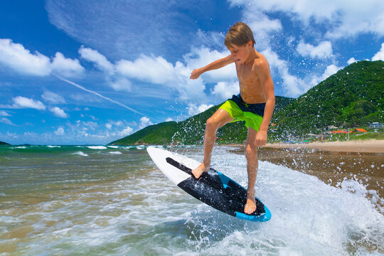 Young boy running, jumping on skimboard and sliding by shallow water on sunny sea beach. Skimboarding, childhood adventure, active lifestyle, watersports, summer vacation and fun outdoor activities.