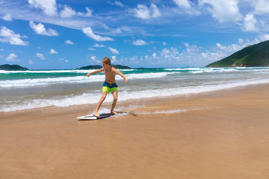 Young boy running, jumping on skimboard and sliding by shallow water on sunny sea beach. Skimboarding, childhood adventure, active lifestyle, watersports, summer vacation and fun outdoor activities.
