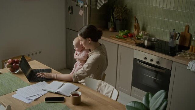 Caucasian young adult mother holding infant while typing on laptop. Clip suitable for advertising remote employment, maternity leave, freelance career, and work-life balance