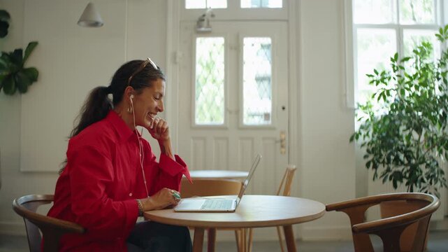 Woman talks with earbuds on a laptop video call in a cafe, reflecting remote work, freelance communication, online meetings, and flexible digital work.