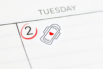 Close up of calendar with sticker showing sanitary pad and red heart. Reminder for tracking menstrual cycle and period start © Firn