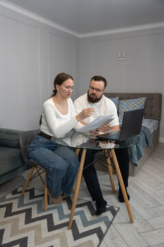 Young couple discussing financial documents, managing personal budget, and making online payments using a laptop in their home living space, focusing on budgeting and debt awareness. Vertical photo