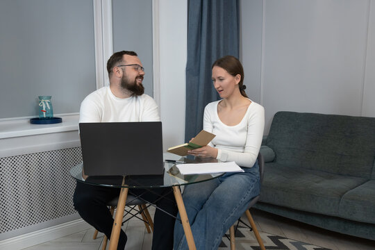 Young couple in a cozy home office sharing ideas: she reads from a book while he watches a laptop, collaborating on a project, smiling and enjoying remote work together