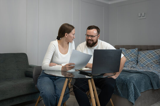 Smiling young couple at home reviewing bills and financial documents on a laptop, planning budget, savings and future investments together in a comfortable living room setting