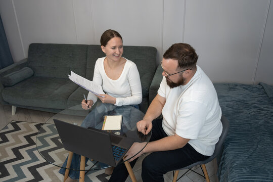 Young adult couple working remotely from home, a man and a woman discussing financial documents and reports near a laptop, planning household budget or future investments