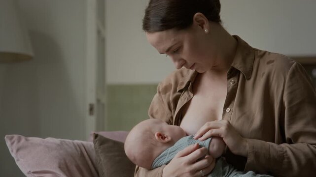 Caucasian young adult woman breastfeeding newborn baby tenderly indoors. Footage suited for maternal healthcare campaigns, infant nutrition promotions, lactation support videos and parenting guides