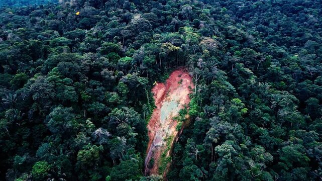 Aerial view of a large landslide scar exposing bare earth on a forested slope surrounded by dense tropical rainforest. The dramatic landscape illustrates soil erosion, slope instability