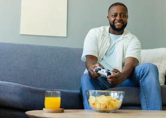 Man sitting on grey sofa in living room, holding white game controller and snacking chips © WavebreakMediaMicro