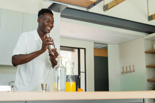 African American man in white shirt shaking cocktail shaker in home kitchen with carafe, jigger