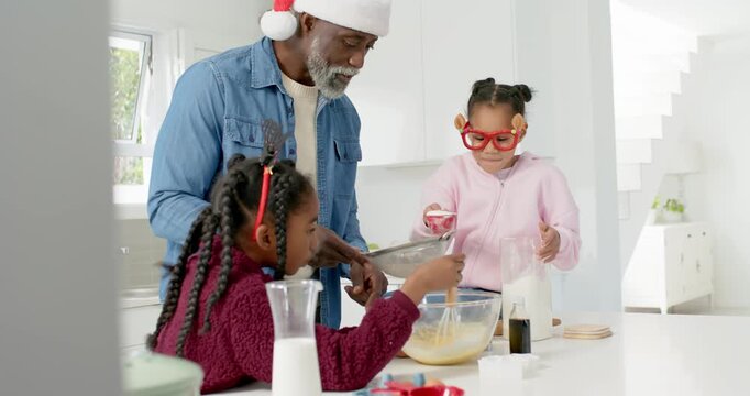 African American dad donning Santa hat guiding two youth girls mixing batter, sifting for christmas