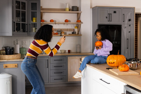 African American female mom taking youth daughter phone pic in kitchen halloween pumpkin