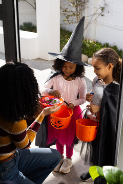 African woman, two female youths offering bowl of candy with pumpkin buckets on porch halloween