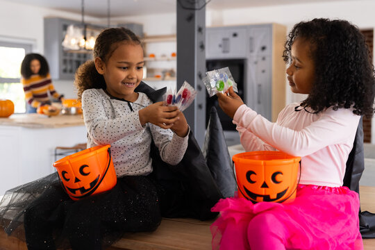 African American female family, two caped kids sharing halloween candies with pumpkins in kitchen