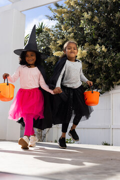 African American female children holding hands hopping on porch in halloween gear with orange pails