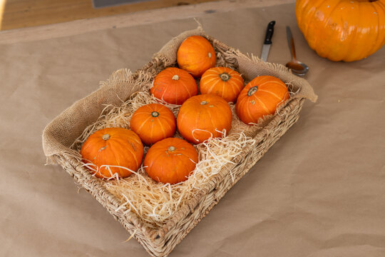 Wicker basket is sitting on brown craft paper, holding nine orange pumpkins, knife, spoon halloween