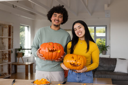 Diverse couple holding carved halloween pumpkins and posing with carving tools on living room table