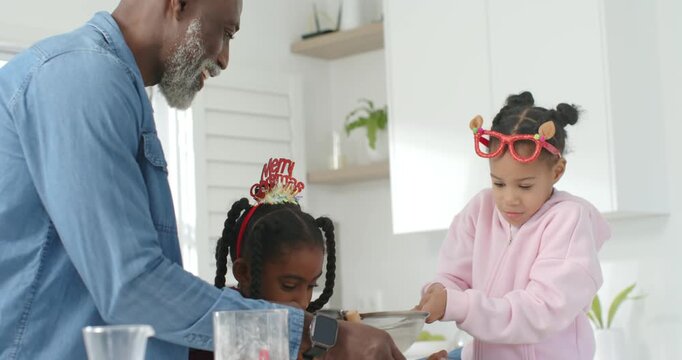African American family guiding two teen girls sifting, whisking bowl in kitchen for christmas