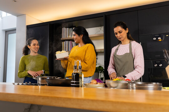 Diverse female friends cooking at wooden kitchen island, holding steaming bowl, chopping produce