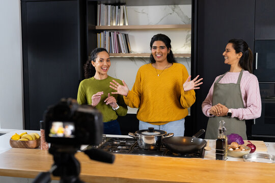 Diverse women hosting cooking demo at kitchen island with camera on tripod and cooktop, olive apron