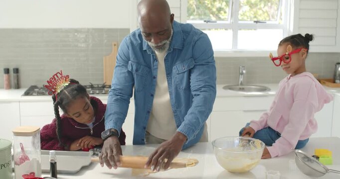 African family watching man rolling dough using pin at island, youth girls by cutters for christmas