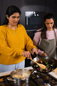 Two women stirring vegetables in wok on gas stove, pot boiling noodles, one in yellow sweater