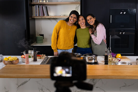 Asian women, one in apron, posing for camera on tripod behind kitchen island, cooking with saucepan