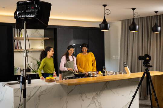 Diverse female team cooking and filming at kitchen island wearing apron, frying pan, camera