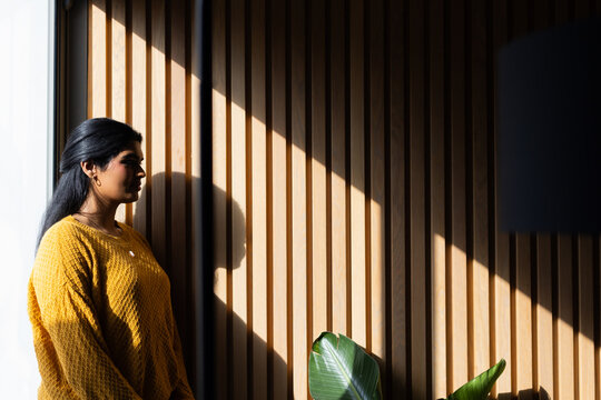 Indian woman standing at home by slat wall wearing mustard sweater, casting shadow, copy space