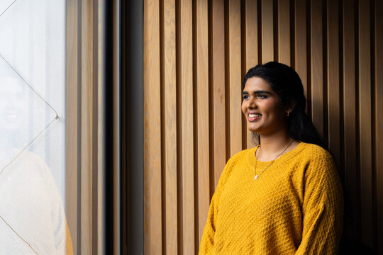 Adult woman posing right by glass window, wearing mustard sweater and pendant, wood slats