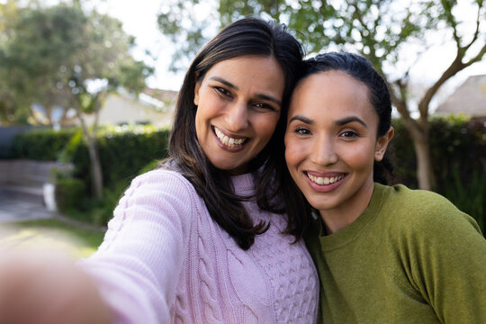 Female friends posing for selfie in garden holding phone and showing pink knit and olive sweaters