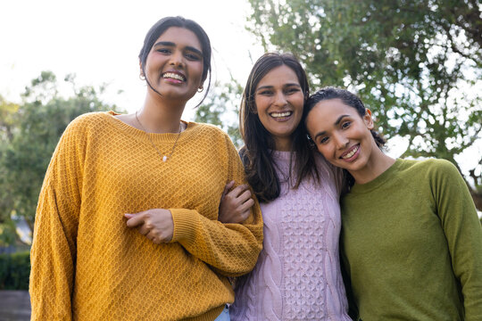 Diverse female friends standing in park linking arms, wearing sweaters and pendant necklace